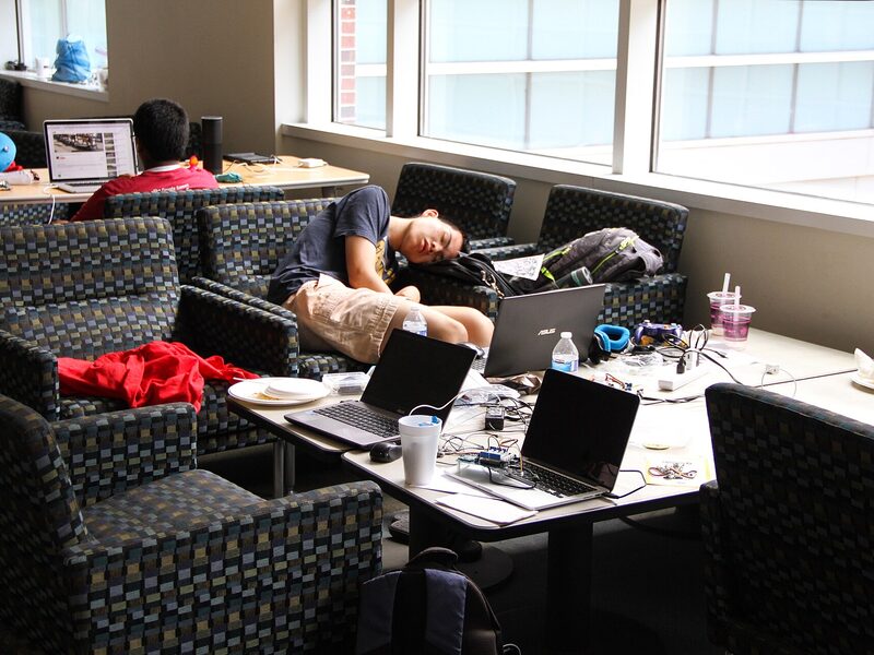 A student asleep on a couch between laptops at a hackathon